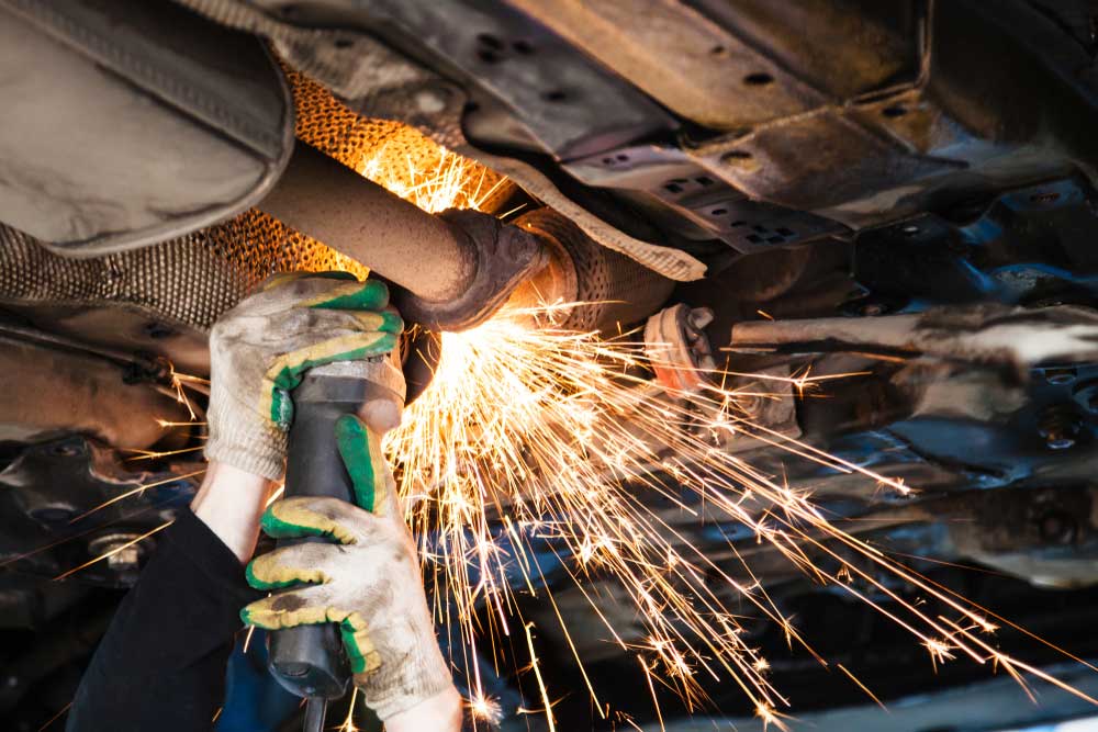 Hands beneath a lifted car using a hand grinder on a muffler